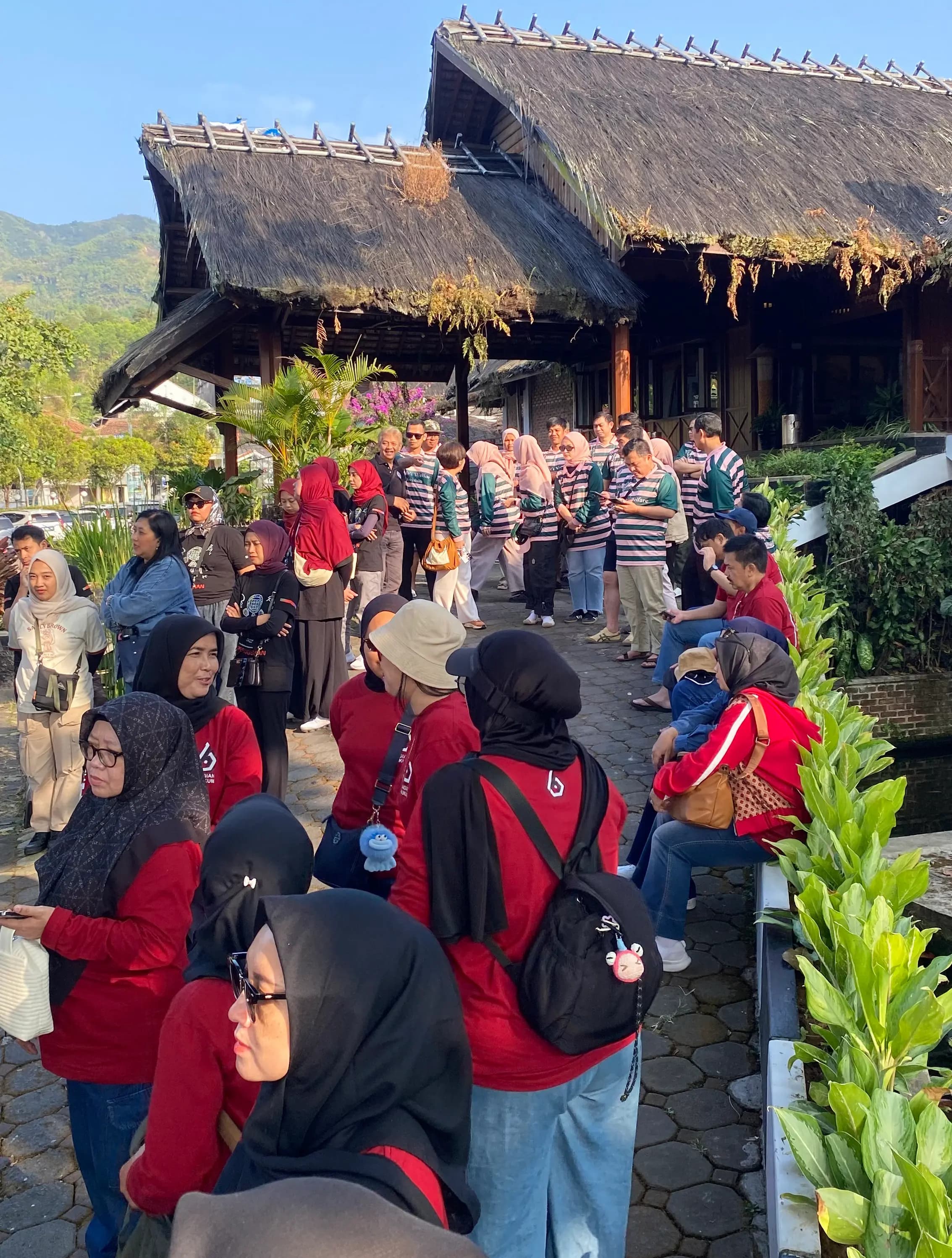 Rombongan tamu berkaus merah berkumpul di halaman basecamp beratap ilalang dengan latar pegunungan